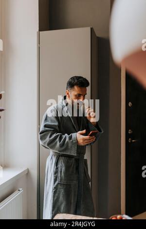 Ein Mann im Bademantel, der sein Smartphone benutzt, während er zu Hause Zähne putzt Stockfoto