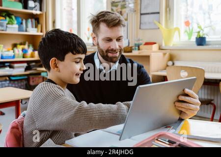 Lächelnder männlicher Lehrer, der dem Schuljungen am Schreibtisch mit einem Tablet-PC hilft Stockfoto