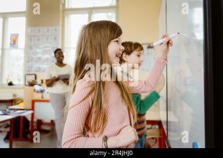 Glückliches Mädchen, das auf dem Whiteboard schreibt, während es Mathematik-Probleme im Klassenzimmer löst Stockfoto