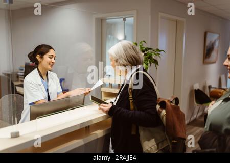 Lächelnder Empfangsmitarbeiter erklärt dem Patienten das Ausfüllverfahren durch durchsichtige Abschirmung in der Klinik Stockfoto