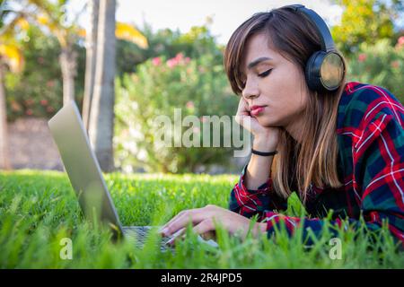 Eine junge Frau benutzt ihren Laptop, während sie auf einer Wiese in einem öffentlichen Park liegt Stockfoto
