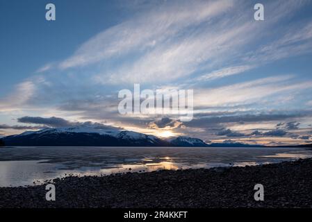 Sonnenuntergang am Rocky Lake in Atlin, British Columbia im Frühling. Schneebedeckte Berge spiegeln sich im ruhigen See mit dramatischem Himmel. Stockfoto