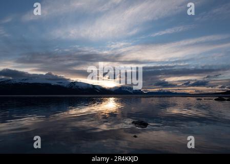 Sonnenuntergang am Rocky Lake in Atlin, British Columbia im Frühling. Schneebedeckte Berge spiegeln sich im ruhigen See mit dramatischem Himmel. Stockfoto