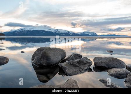 Sonnenuntergang am Rocky Lake in Atlin, British Columbia im Frühling. Schneebedeckte Berge spiegeln sich im ruhigen See mit dramatischem Himmel. Stockfoto