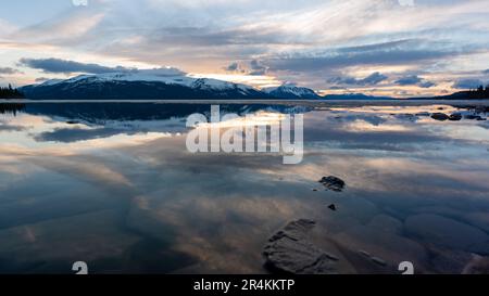 Sonnenuntergang am Rocky Lake in Atlin, British Columbia im Frühling. Schneebedeckte Berge spiegeln sich im ruhigen See mit dramatischem Himmel. Stockfoto