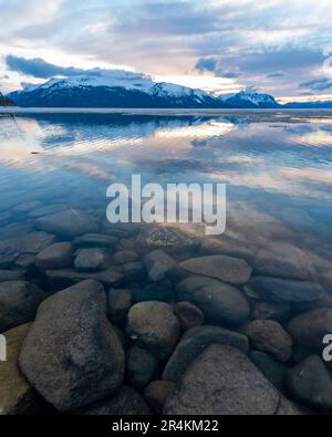 Sonnenuntergang am Rocky Lake in Atlin, British Columbia im Frühling. Schneebedeckte Berge spiegeln sich im ruhigen See mit dramatischem Himmel. Stockfoto