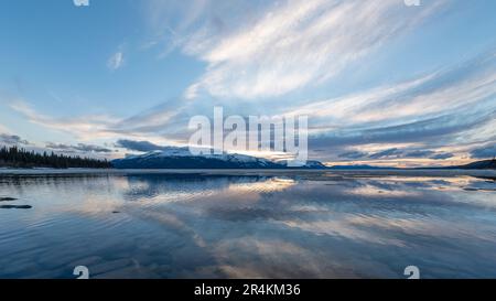 Sonnenuntergang am Rocky Lake in Atlin, British Columbia im Frühling. Schneebedeckte Berge spiegeln sich im ruhigen See mit dramatischem Himmel. Stockfoto