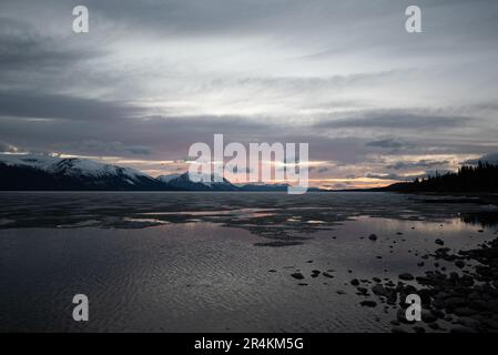 Sonnenuntergang am Rocky Lake in Atlin, British Columbia im Frühling. Schneebedeckte Berge spiegeln sich im ruhigen See mit dramatischem Himmel. Stockfoto