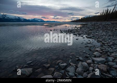 Sonnenuntergang am Rocky Lake in Atlin, British Columbia im Frühling. Schneebedeckte Berge spiegeln sich im ruhigen See mit dramatischem Himmel. Stockfoto