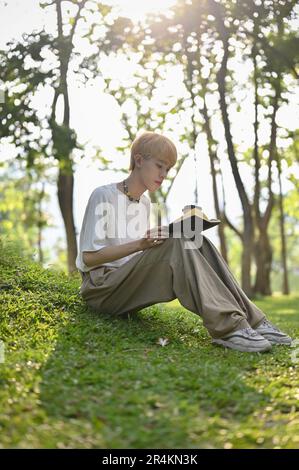 Das Porträt eines jungen asiatischen Schwulen konzentriert sich darauf, ein Buch zu lesen, während man sich an einem sonnigen Tag allein im grünen Park entspannt. LGBTQ und Lifestyle-Konzepte Stockfoto