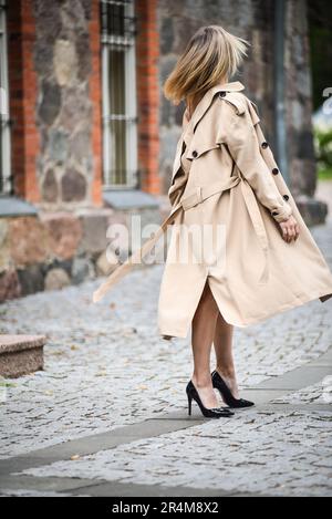 Eine stilvolle Frau im beigen Regenmantel mit modischer Frisur geht durch die Straßen der Stadt Stockfoto