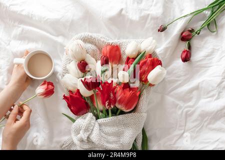 Ein Blumenstrauß und eine Tasse Kaffee in weiblichen Händen in einem weißen Bett. Stockfoto