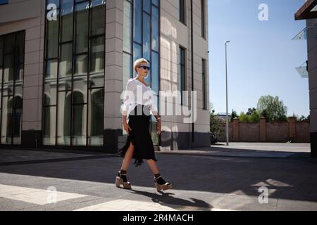 Selbstvertrauen und wunderschöne blonde Frau mit stilvollem, kurzen Haarschnitt wandern durch die Straßen der Stadt in einem schicken weißen Hemd und einem schwarzen Rock mit Fransen und hohem Oberteil Stockfoto