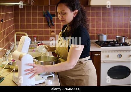 Schöne schwangere Hausfrau mit beigefarbener Küchenschürze, die mit einer elektrischen Küchenmaschine Sahne schlägt Stockfoto