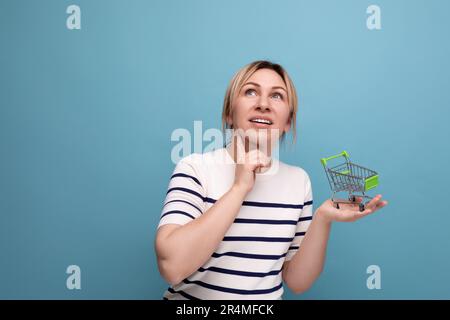 Ein blondes, blondes, attraktives Mädchen in einer lässigen Jacke denkt darüber nach, einen Einkaufswagen in der Hand zu halten, auf blauem Hintergrund Stockfoto