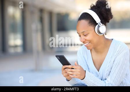 Glückliche schwarze Frau, die auf der Straße mit dem Smartphone Musik mit Kopfhörern hört Stockfoto