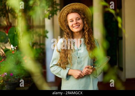 Junge Gärtnerin mit Strohhut, die die Handschaufel hält, steht im Garten und sieht die Kamera an und lächelt fröhlich. Fröhliche Weiße mit grünem Obstgarten Stockfoto