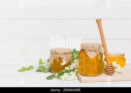 Süßes Honigglas umgab die Akazienblüten des Frühlings. Honig fließt aus einem Löffel in ein Glas. Gläser von klarem frischen Akazienhonig auf Holzgrund. Stockfoto