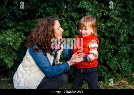 Eine lachende Mutter und ein kleines Kind sitzen zusammen vor dem beleuchteten Baum Stockfoto