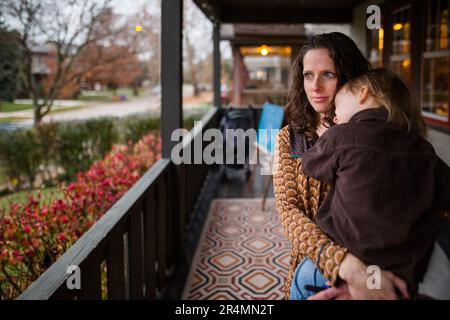 Eine mutter steht auf der Veranda, hält ein kleines Kind und schaut auf die Straße Stockfoto