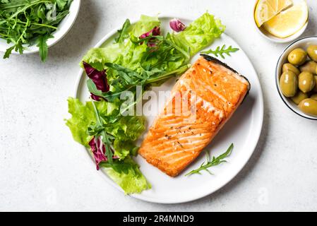 Lachssteak und frischer Blattgemüsesalat mit Rucola und Oliven auf weißem Hintergrund. Mediterranes Keto Paleo Essen - grüner Salat und geröstet Stockfoto