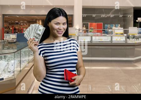 Lächelnde junge Asiatin zeigt Fan von Dollars aus der roten Brieftasche. Innenausstattung des Einkaufszentrums im Hintergrund. Stockfoto