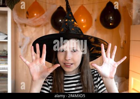 Kleine hübsche Hexe mit schwarzem Zauberhut sagt Buh mit beängstigender Geste. Halloween-Kinder-Porträt. Mädchen im Halloween-Kostüm zu Hause. Das Kind ist angezogen Stockfoto