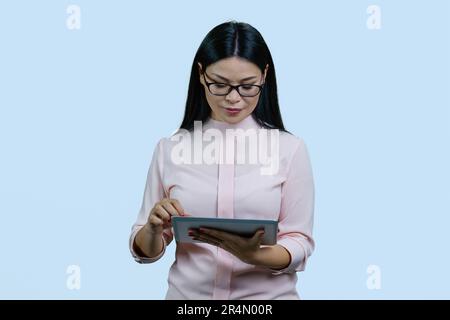 Eine junge, intelligente Asiatin mit Brille verwendet einen Tablet-pc. Nach unten schauen und scrollen. Isoliert auf blassblauem Hintergrund. Stockfoto