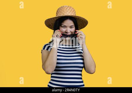 Eine junge asiatische Touristenfrau mit Strohhut zieht eine Sonnenbrille an. Isoliert auf gelbem Hintergrund. Stockfoto