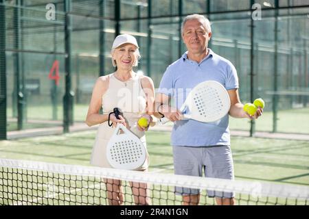Alter Mann und Frau posieren mit Padel Schläger in den Händen neben dem Netz auf dem Tennisplatz Stockfoto