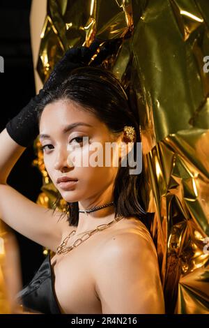 Elegante junge asiatische Frau mit nasser Frisur und kurzen Haaren in trägerlosem Kleid mit schwarzem Handschuh, während sie neben goldenem Hintergrund steht, Model Stockfoto
