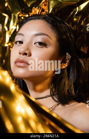 Porträt einer wunderschönen jungen asiatischen Frau mit nassem kurzen Haar, die neben glänzendem goldenen Hintergrund posiert, Model, in die Kamera schaut, gelbes Faltenblatt, na Stockfoto