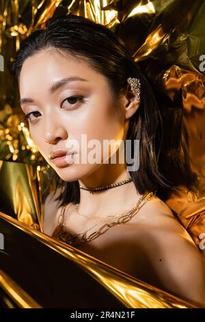 Porträt einer wunderschönen jungen asiatischen Frau mit nassen kurzen Haaren, die neben glänzendem Hintergrund posiert, Model, in die Kamera schaut, goldene Faltenfolie, natürliches m Stockfoto
