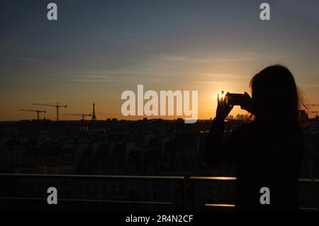 Paris bei Sonnenuntergang. Eine junge Frau fotografiert mit ihrem Handy die Pariser Stadtlandschaft mit Eiffelturm. Blick vom Centre Pompidou. Stockfoto