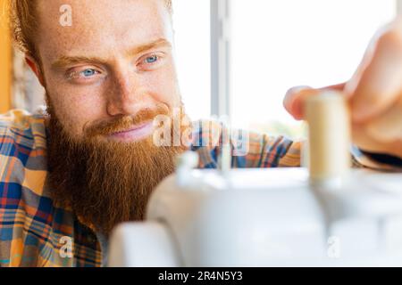 Hübscher rothaariger Mann mit langen Bartnähten an einer Nähmaschine im Studio Stockfoto