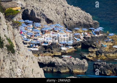 Playa rocosa entre acantilados, desde arriba, llena de sombrillas blancas y azules, en la isla de Capri Stockfoto