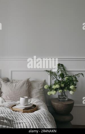 Frühstück im Bett. Eine Tasse Kaffee, ein Korbblech. Strauß aus weißem Viburnum, Farn und salomonen Robbenblüten. Moderner Bo-Ho-Nachttisch. Zimmer mit Blick auf das Schlafzimmer. Beige Stockfoto