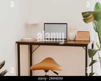 Stilvolle Inneneinrichtung im Home Office mit Laptop-Bildschirm-Mockup, Holztisch, Pflanze, Notizen, Sattelhocker und elegantem Bürozubehör im Designer-Apartment. Stockfoto