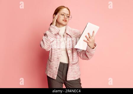 Träumende junge Geschäftsfrau, eine Schülerin in stilvollem Kostüm und eine Brille, die ein Tablet über einem pinkfarbenen Studiohintergrund hält. Bildungskonzept, online Stockfoto