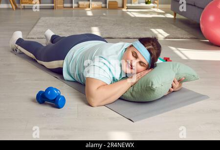 Lustige glückliche faule, fette Frau, die auf Kissen und Sportmatte schläft, anstatt Fitnesstraining zu machen Stockfoto