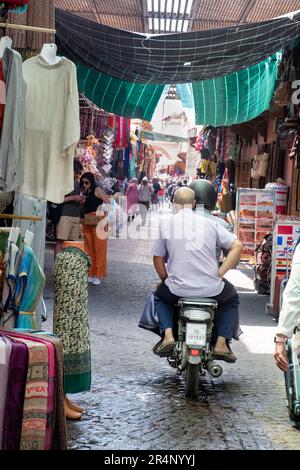 Ein Motorrad, das durch eine enge Gasse zwischen den Souks gefahren wird, mit vielen Einkäufern und Fußgängern im zentralen Souk-Gebiet von Marrakesch marokko. Stockfoto