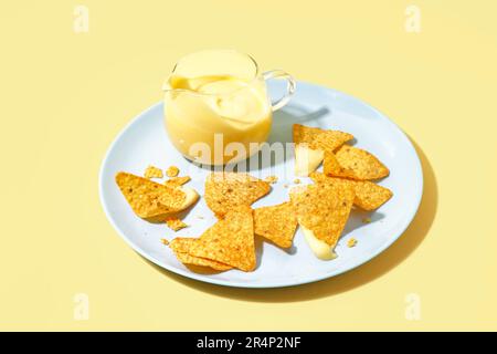 Teller mit Krug mit schmackhafter Cheddar-Käsesauce und Nachos auf blassgelbem Hintergrund Stockfoto
