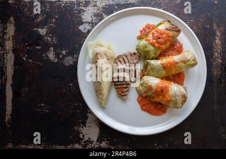 Kohlbrötchen mit saftig gegrilltem Schweinefilet, serviert neben gekochtem Kohl und einer lebhaften Tomatensoße, elegant auf makellosem Boden angeordnet Stockfoto