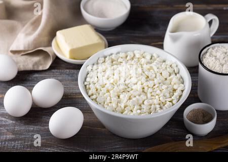 Schüssel mit Hüttenkäse, Eiern, Mehl, Butter, Milch, Zucker, Vanillezucker auf Holzhintergrund. Zutaten für das Backen zu Hause Stockfoto
