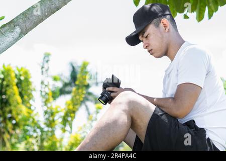 lateinamerikanischer Influencer, der auf dem Gras unter einem Baum sitzt und sich die Bilder ansieht, die er mit seiner Kamera gemacht hat Stockfoto