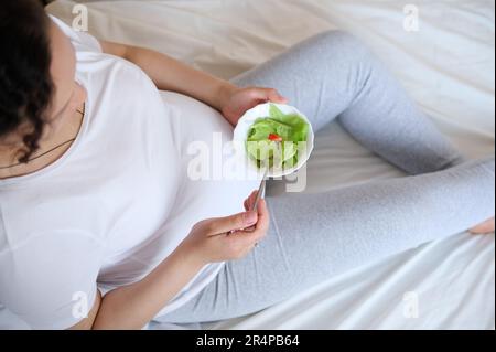 Blick von oben auf eine schwangere Frau, die eine Schüssel mit frischem Salat hält und während der Schwangerschaft einen gesunden Lebensstil genießt Stockfoto