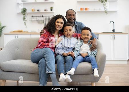Porträt einer multiethnischen vier-Personen-Familie, die an sonnigen Tagen auf einer weichen Couch in einer offenen Küche posiert. Fröhliche Mutter und süße Söhne kuscheln sich in Vaters Arme und genießen Komfort und Sicherheit. Stockfoto