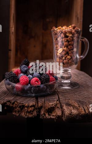 Verschiedene Beeren auf einer Glasplatte. Schokoladengranola in einem Glas. Zusammensetzung auf dem alten Vorstand und auf dem Hintergrund der Box. Vertikaler Rahmen. Stockfoto