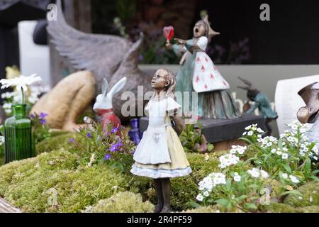 Eine Landszene von Alice in Wonder bei der Chelsea Flower Show, Alice mit Blick nach oben und die Herzkönigin im Hintergrund mit einem Glas Wein. Stockfoto