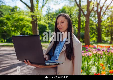 Außenporträt einer jungen Geschäftsfrau, die im Spring Park auf einem Laptop arbeitet. Landschaftsdesignerin genießt die Ergebnisse blühender Blumenbeete in der Öffentlichkeit Stockfoto
